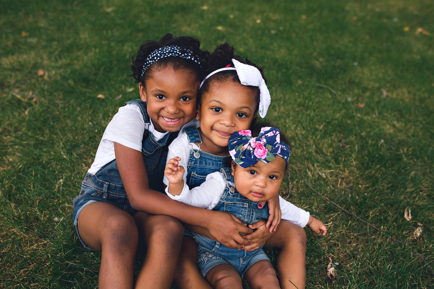 Three adorable sisters take a picture in the grass wearing jean overalls and a white t-shirt. Aren't they just one adorable family?