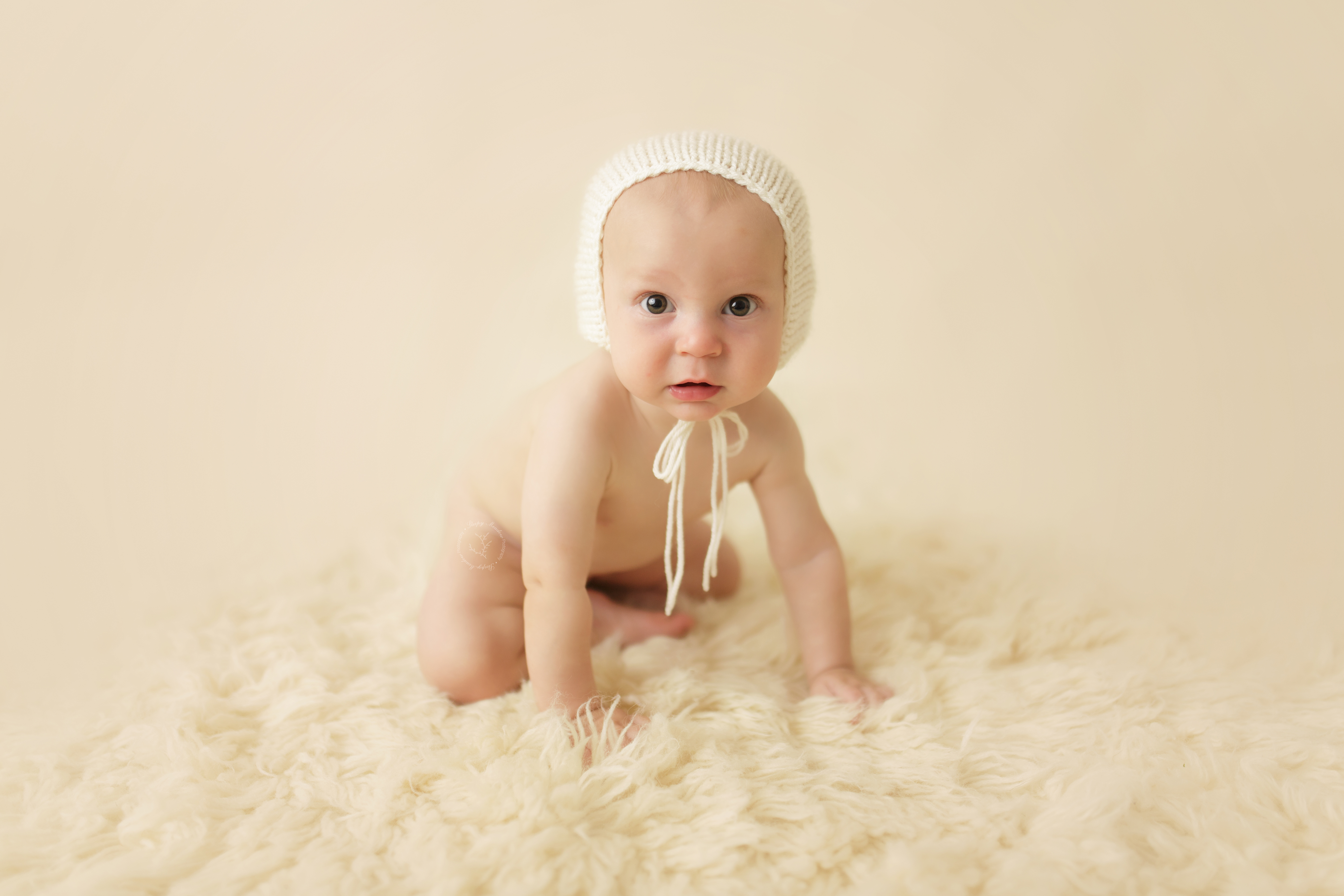 Cute baby boy photographed on cream flokati rug wearing a cream colored knit bonnet.
